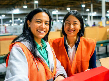 Zwei Frauen in orangefarbenen Westen lächeln in einer Lagerhalle Zwei Frauen in orangefarbenen Westen lächeln in einer Lagerhalle
